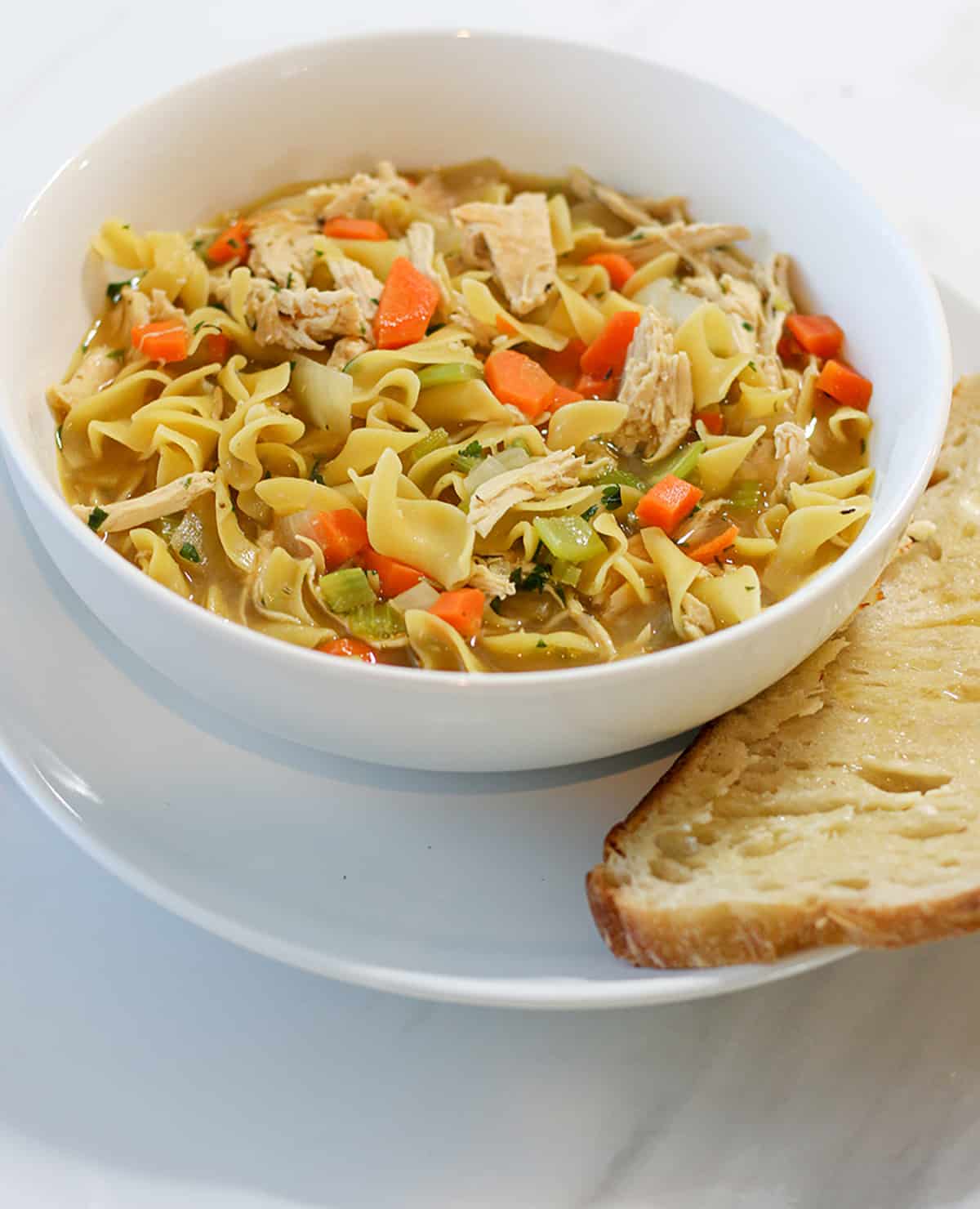 A white bowl with chicken noodle soup and side of sourdough bread.