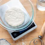 A digital scale with a bowl of sourdough starter, dough whisk, flour and a cup of water.