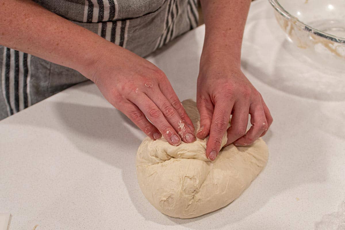 Two hands gently folding over each corner of a loaf of sourdough during the shaping phase.