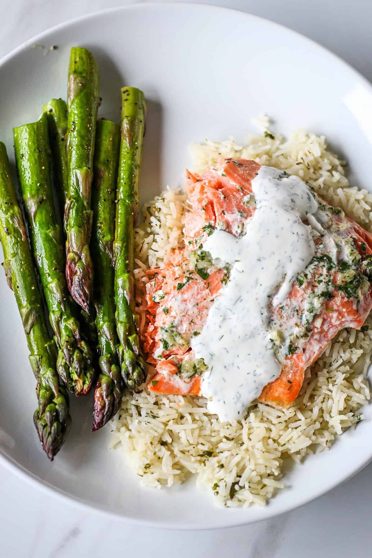 A white plate with salmon asparagus and rice, topped with a sauce and herbs. 