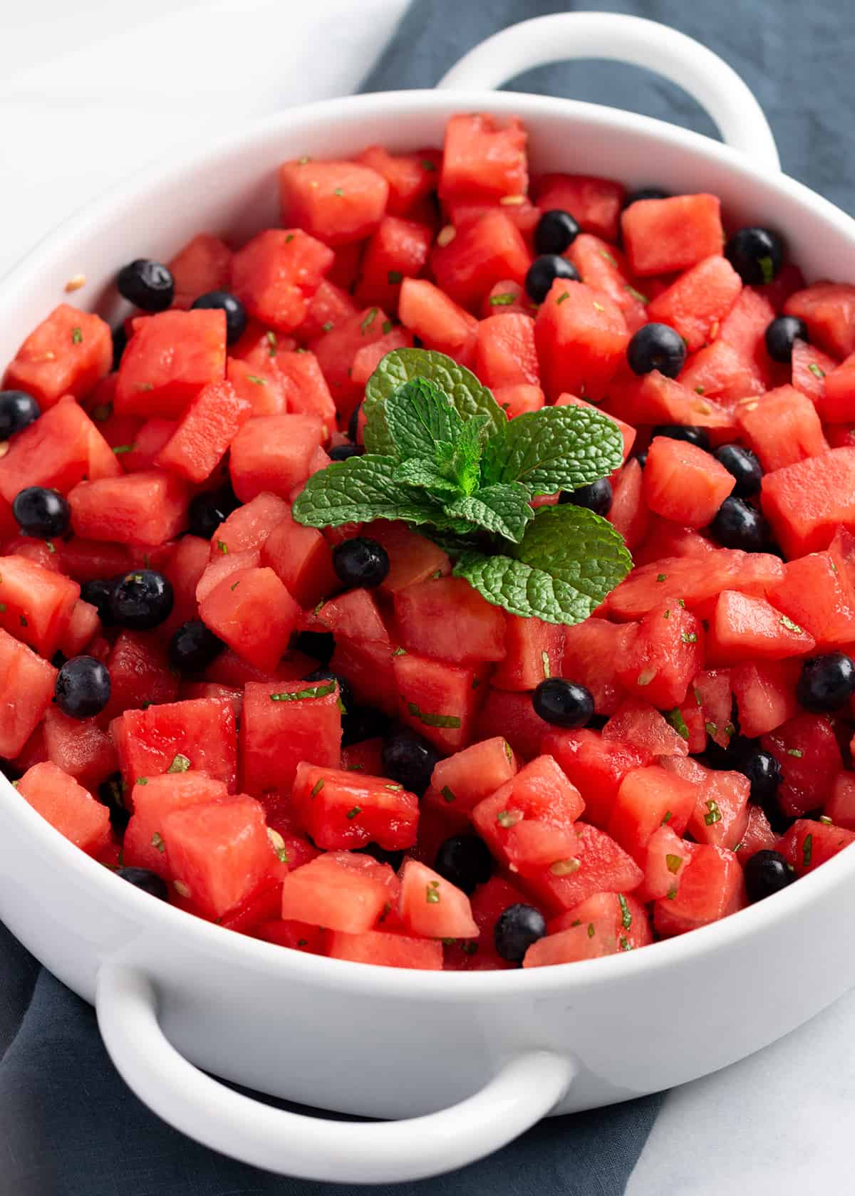 A white serving platter on a blue cloth napkin, filled with fresh watermelon, blueberries and a spring of mint on top. 