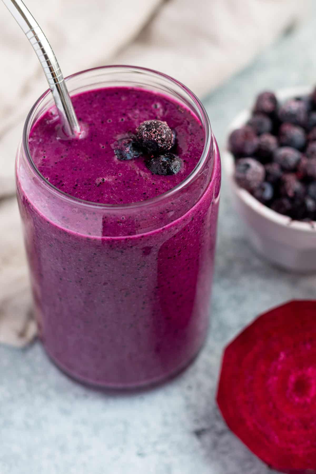 A glass cup with a purple smoothie and metal straw and a raw beet and frozen blueberries