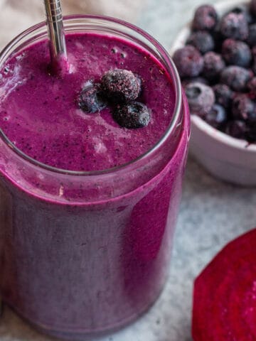 Glass cup with a purple smoothie, metal straw and frozen blueberries.