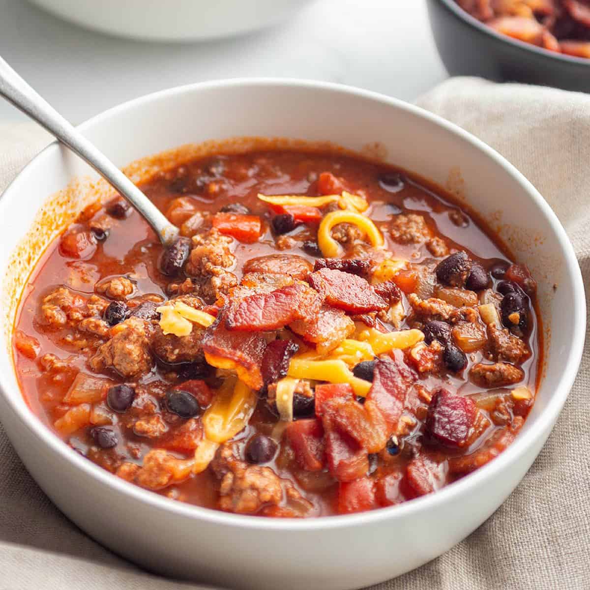 A white bowl with a spoon taking a scoop out of a chili with bacon and cheese.