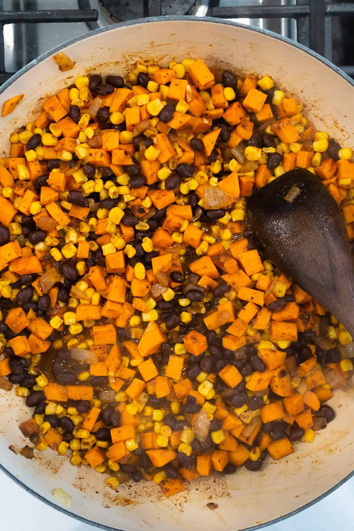 Enamel cast iron with sweet potatoes, corn, black beans being stirred by a wooden spoon.