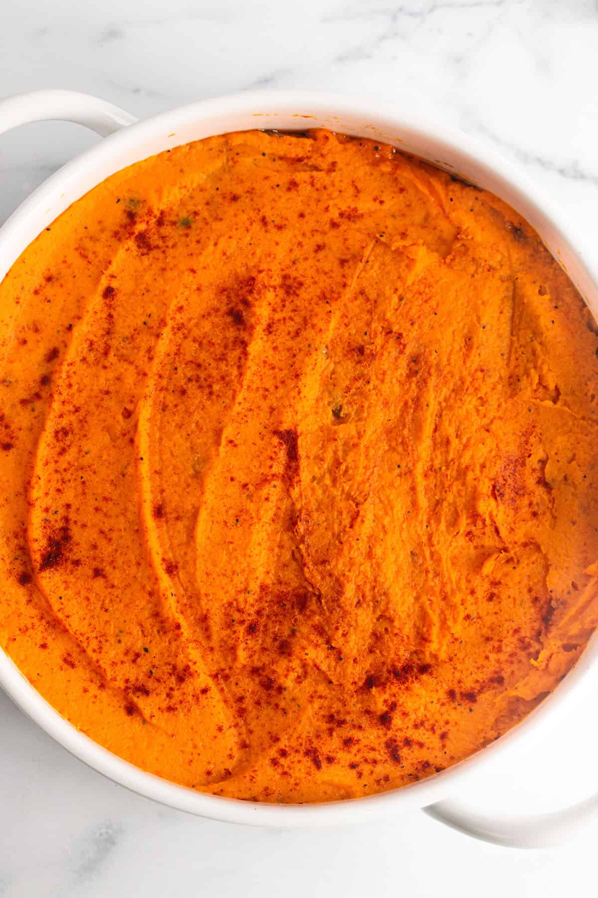 A white circular baking dish with mashed sweet potatoes and topped with paprika.