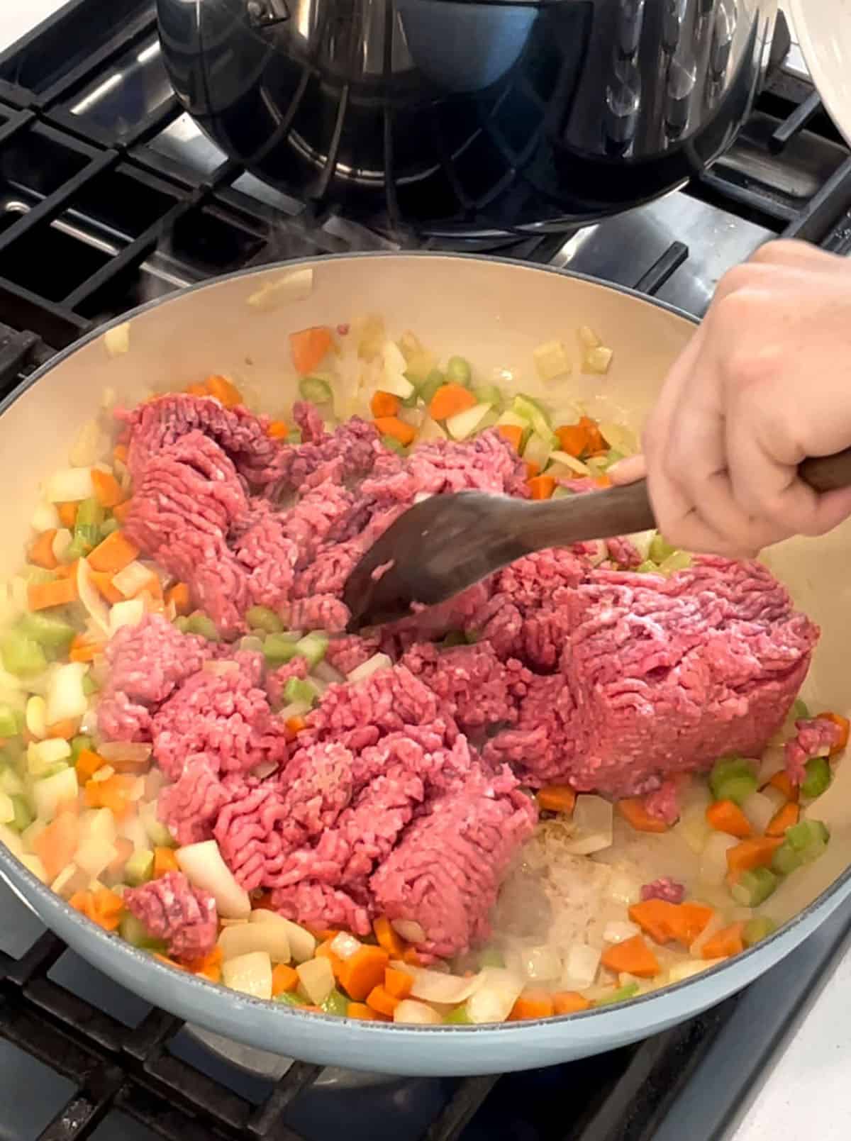 Large skillet with sautéed veggies and raw ground meat being added with a wooden spoon.