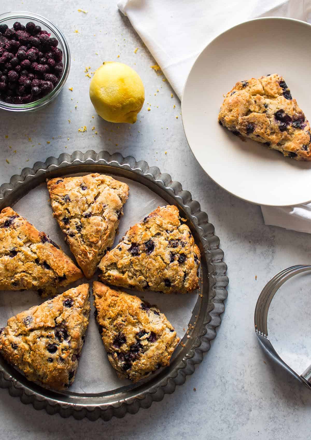 A tart tin with 5 blueberry lemon scones on it. Frozen blueberries, a lemon and a plate with a single scone on it. 