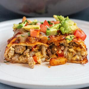 A plate with a breakfast enchilada cut in half with potatoes and sausage topped with avocado and tomatoes.