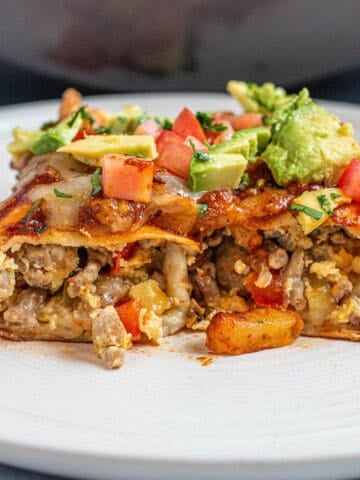 A plate with a breakfast enchilada cut in half with potatoes and sausage topped with avocado and tomatoes.