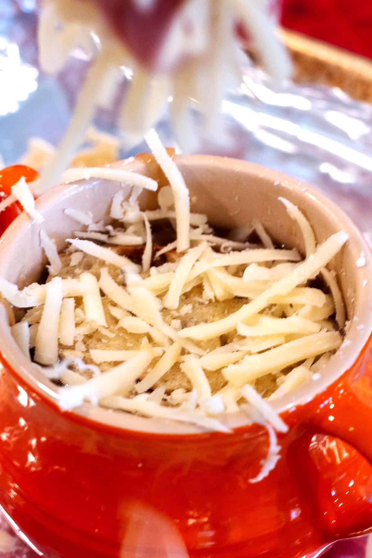 A small red soup pot with grated Swiss cheese being sprinkled on top.