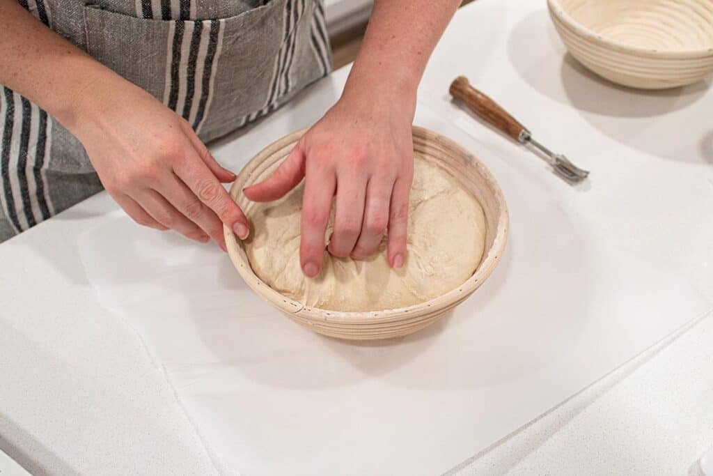 Two hands gently preparing to remove bread dough from a banneton basket.
