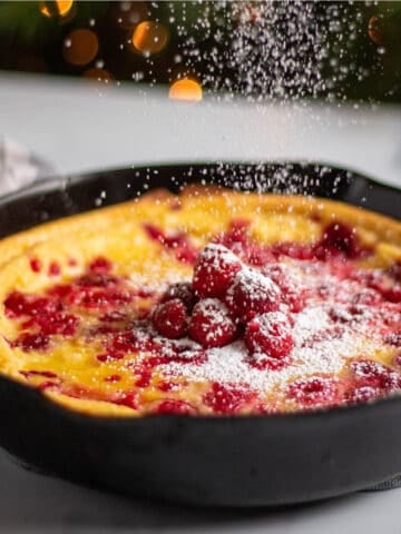 A cast iron pan with a dutch baby filled with raspberries and powdered sugar being dusted on top.