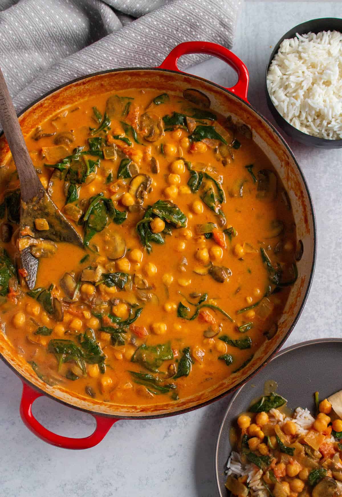 A braiser with spinach and chickpea curry with a bowl of rice and a wooden spoon.