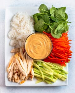 A plate with spinach, noodles, carrots, peppers, cucumbers, chicken and a bowl of peanut sauce in the middle.