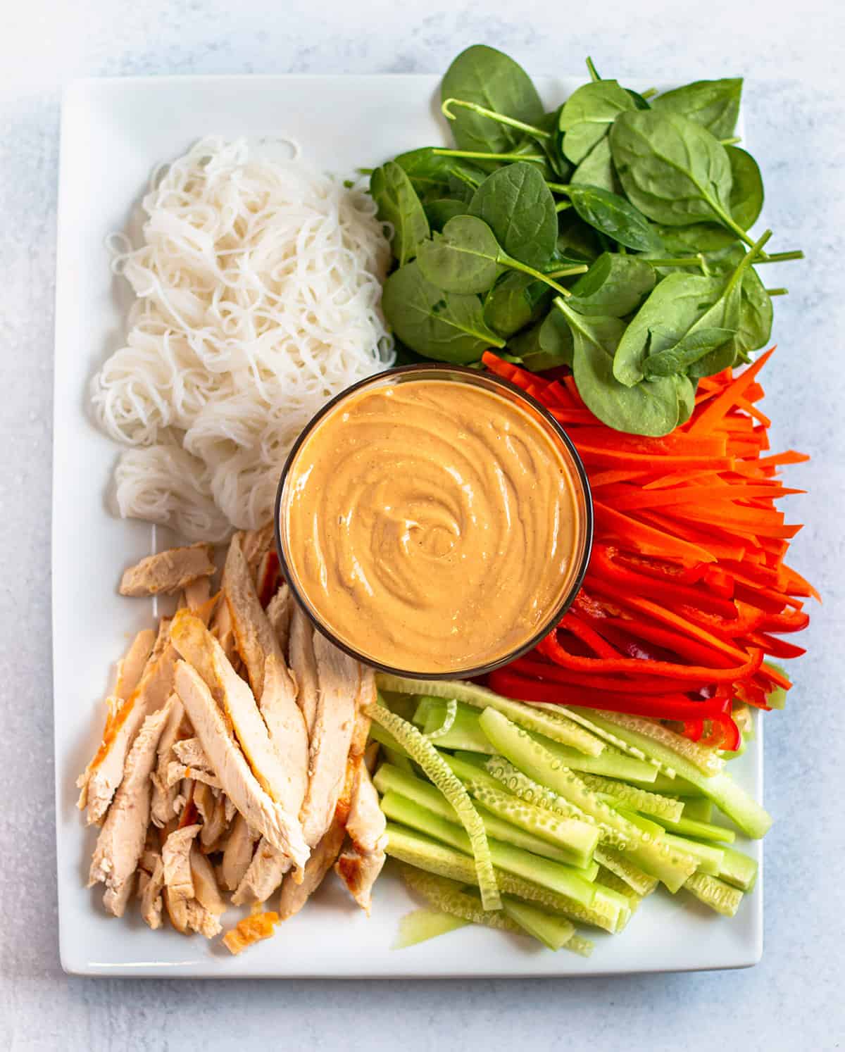 A plate with vermicelli noodles, baby spinach, carrots, red bell peppers, cucumbers chicken and peanut sauce. 