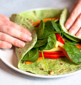 Two hands folding over each side of a spinach wrap over the top of the toppings.