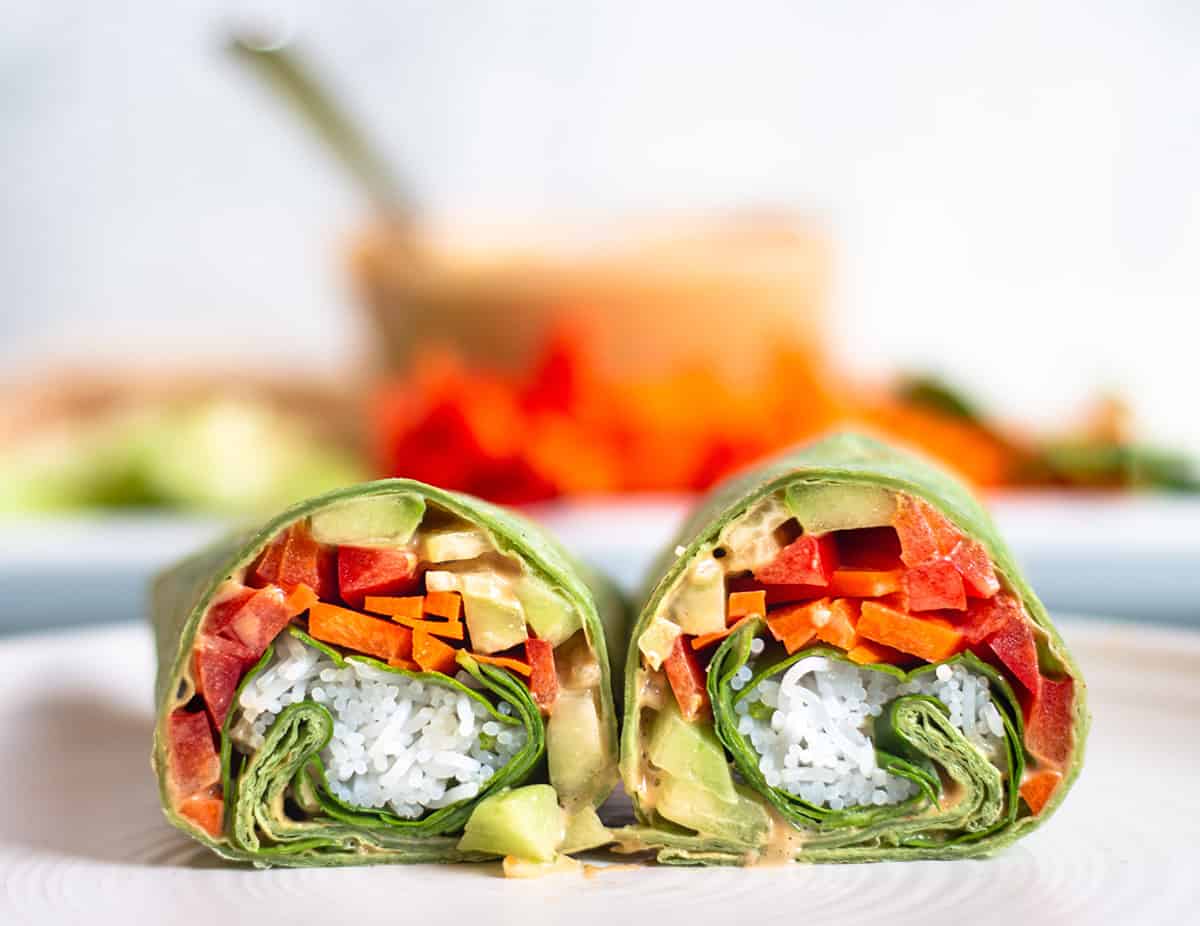 A spinach wrap with fresh veggies, chicken, rice noodles, and peanut sauce cut in half on a plate.