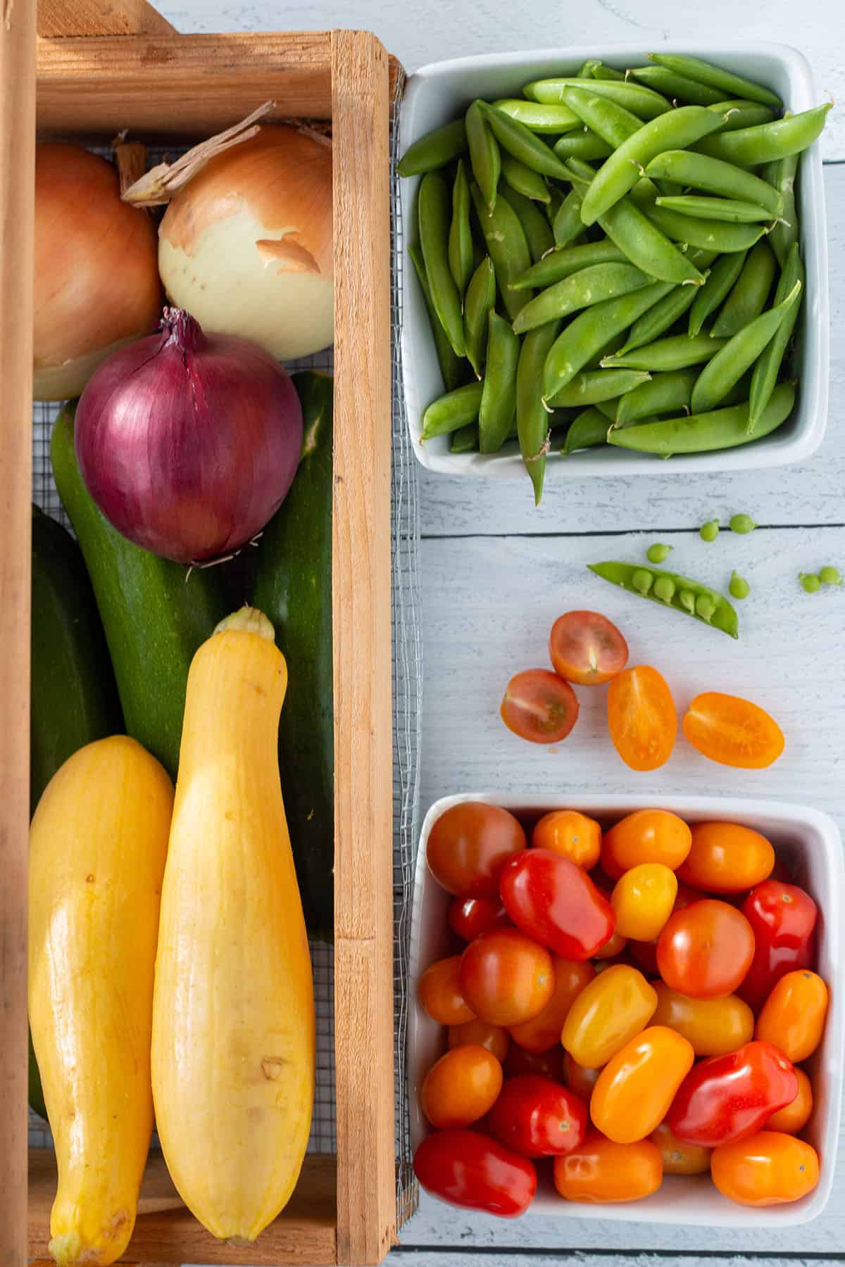 A gardening basket full of squash and onions, with baskets of tomatoes and sugar snap peas.