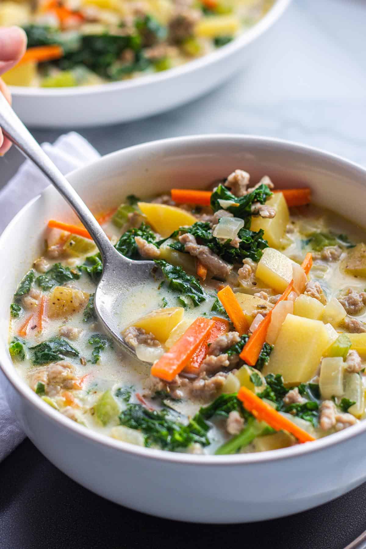 A bowl with a spoon full of soup with potatoes, sausage, veggies and kale. 