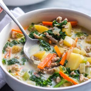 A bowl of soup that is loaded with veggies, potatoes and sausage with a spoon taking a scoop out.