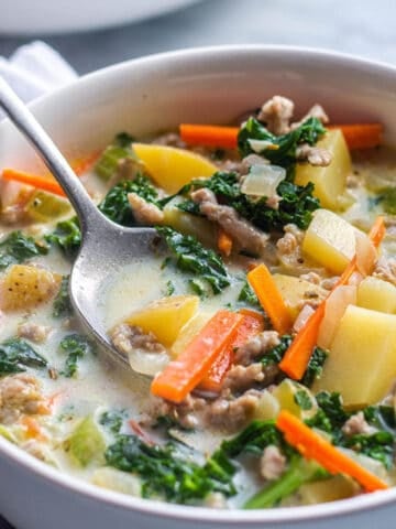 A bowl of soup that is loaded with veggies, potatoes and sausage with a spoon taking a scoop out.