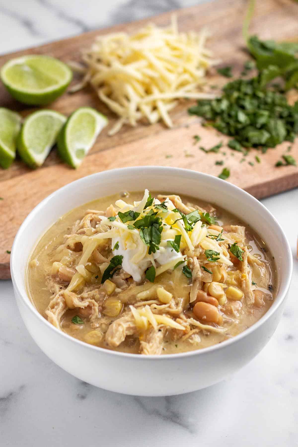 Bowl with white chicken chili soup topped with sour cream, cilantro and cheese. In the back is limes, shredded cheese and cilantro on a board. 