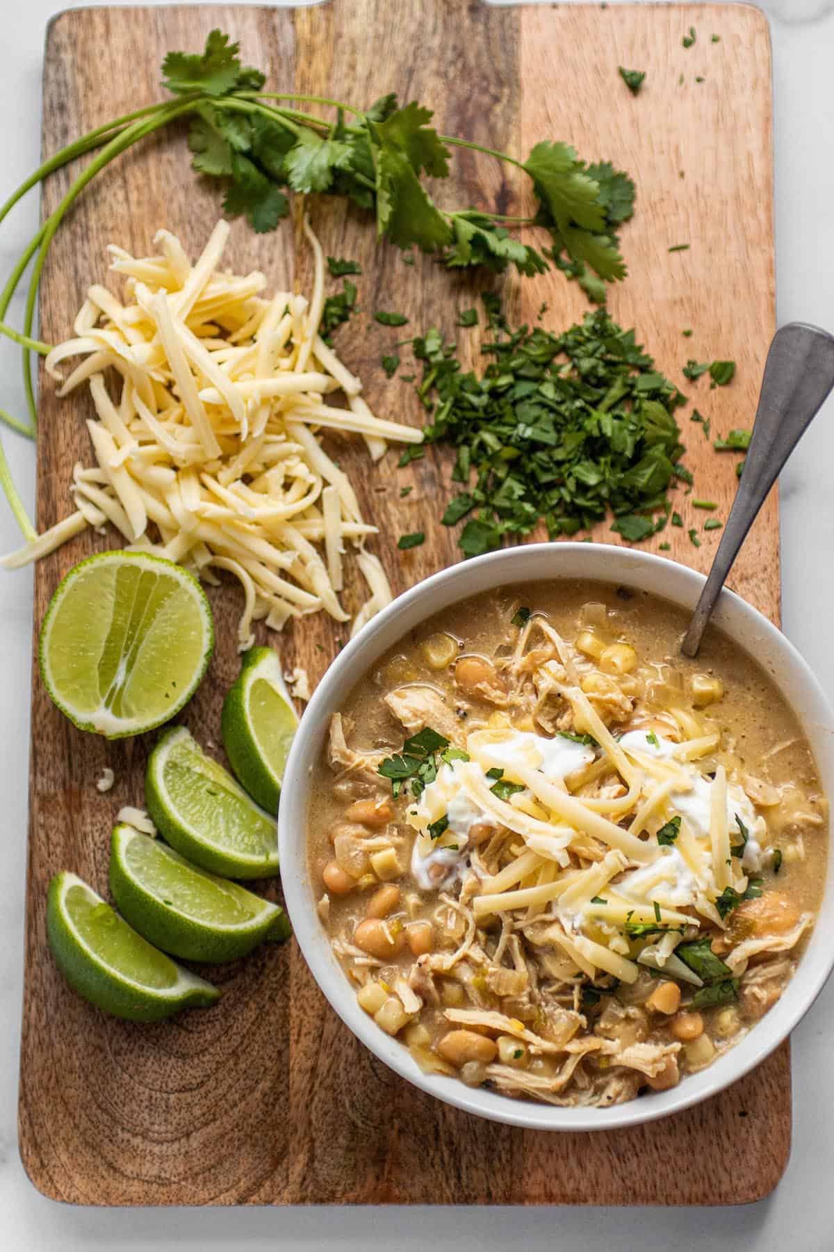 Serving board with a bowl of white chicken chili, limes, shredded cheese and cilantro. 