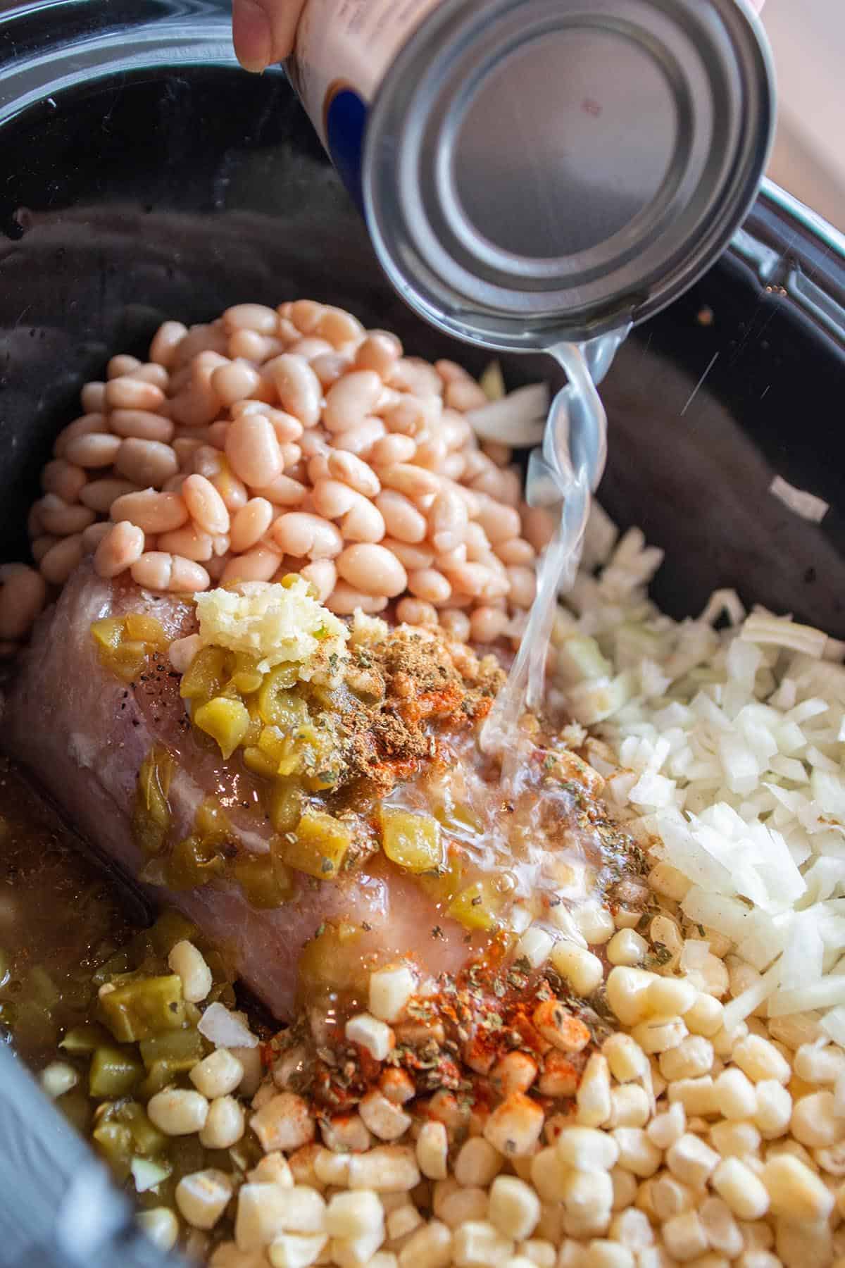 Crock-pot full of fresh onions, white beans, corn, frozen chicken and spices with chicken broth being poured in. 