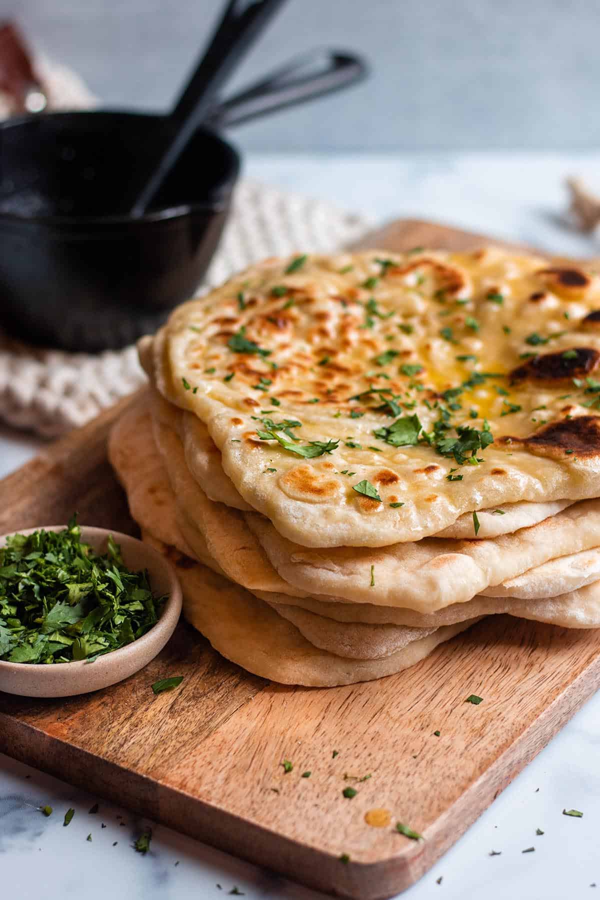 A stack of sourdough naan on a wooden board with cilantro and melted butter. Bowl with fresh cilantro and cast iron butter warmer. 