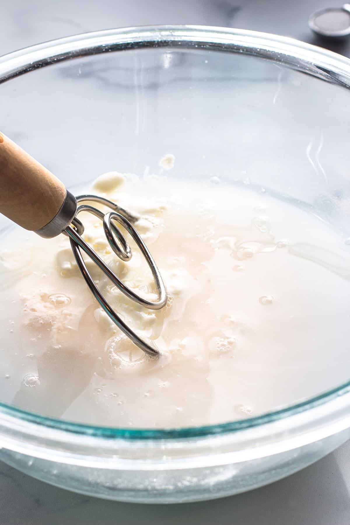 A glass bowl with sourdough discard, yogurt and water being mixed with a dough whisk. 