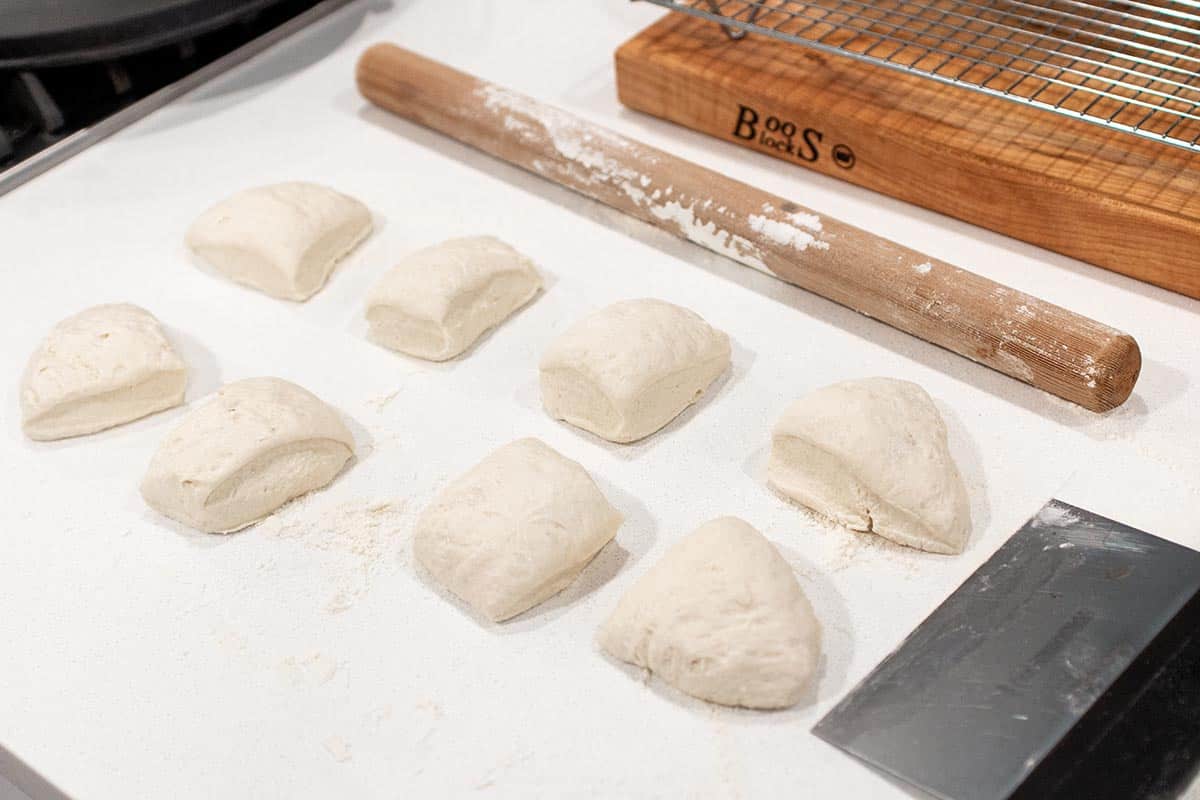 A loaf of dough divided into 8 pieces getting ready to be rolled out. With a bench scraper and rolling pin. 