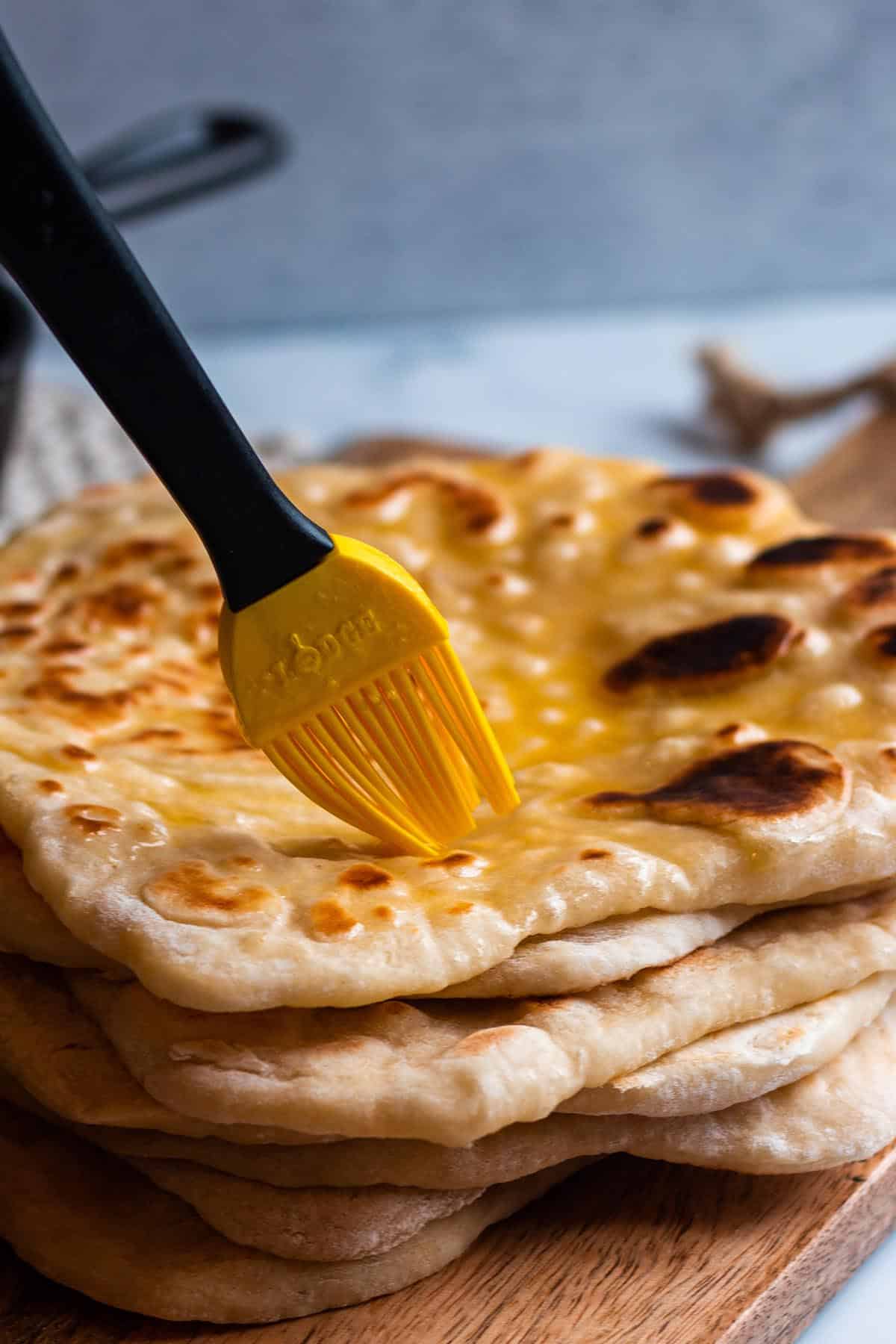 A stack of freshly cooked sourdough naan with a brush spreading on melted butter.