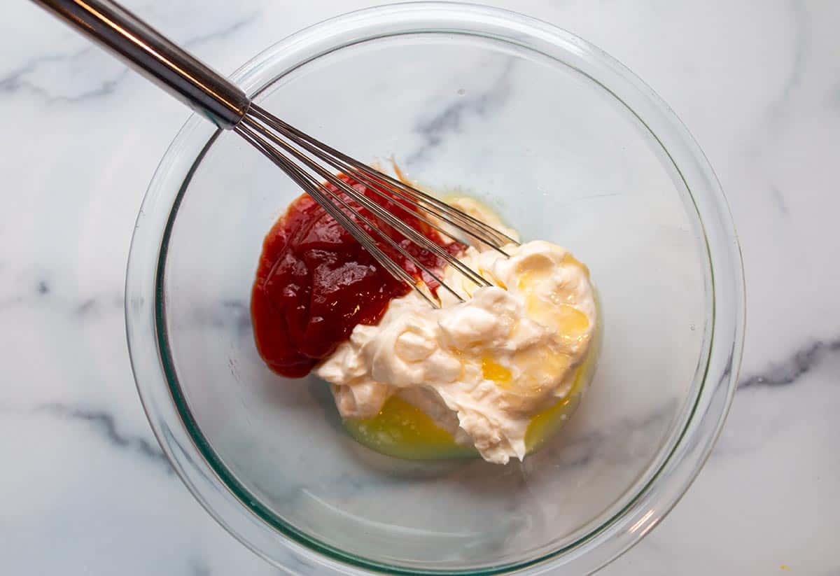Glass bowl and whisk with mayo, melted butter, and ketchup. 