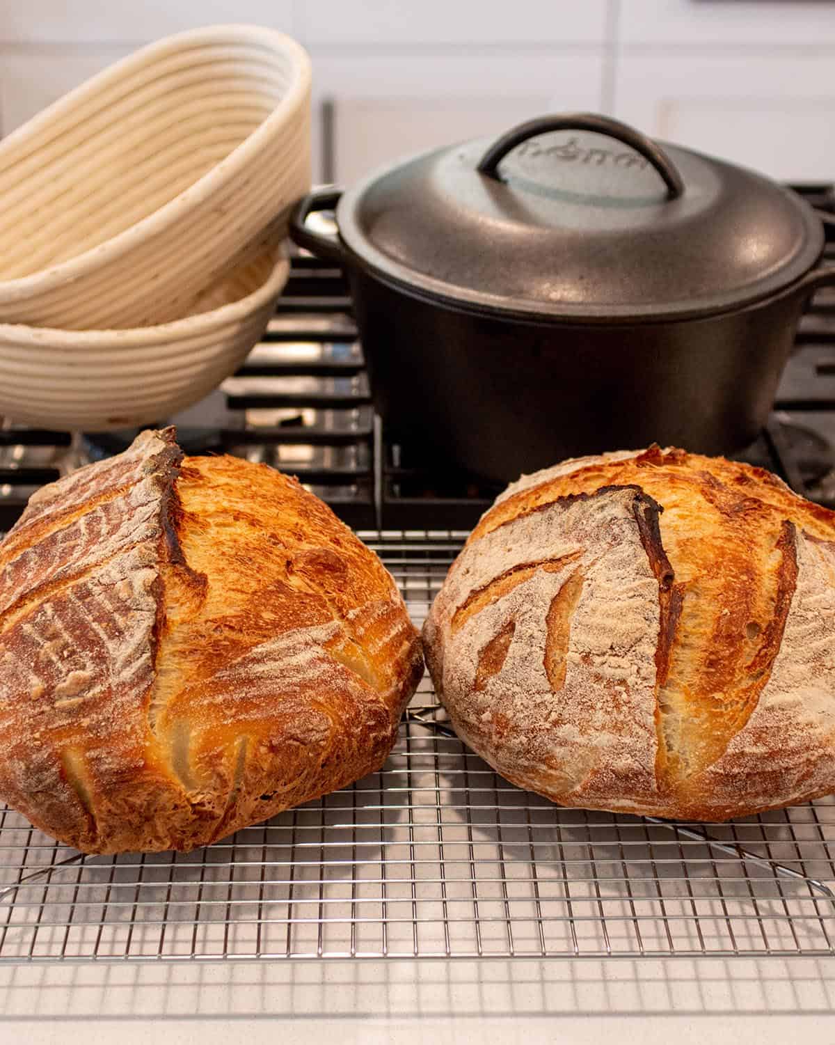 Two loaves of sourdough bread on a cooling rack with two banneton baskets and a cast iron dutch oven. 