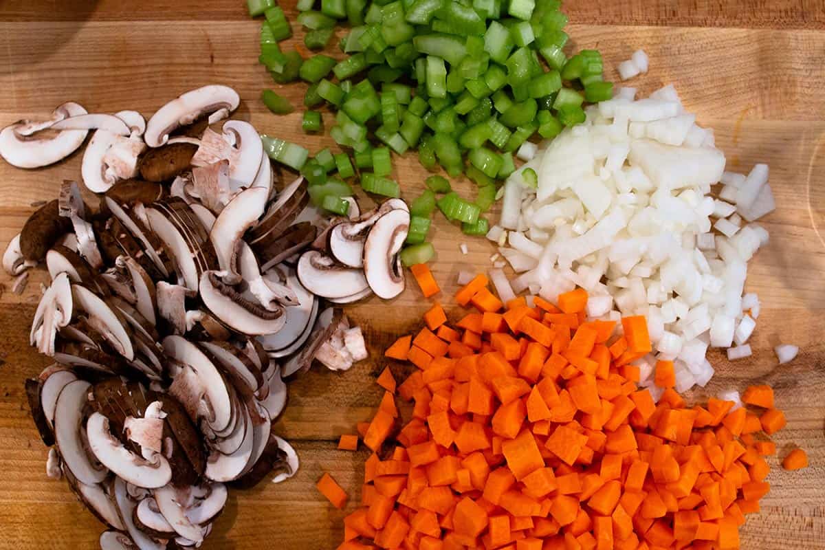Chopping block with cut up celery, carrots, onions and baby bella mushrooms.