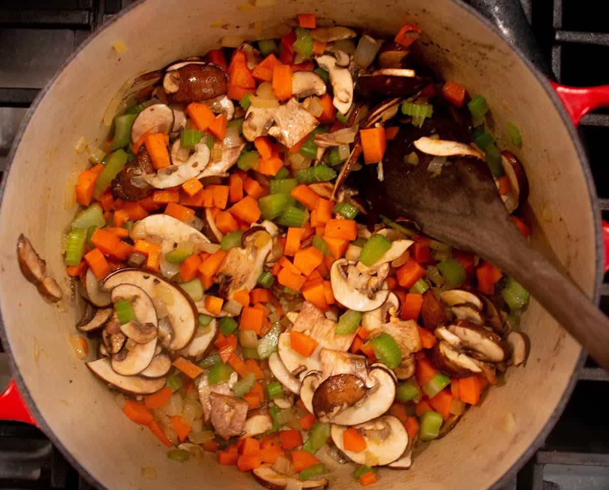 Dutch oven with wooden spoon stirring mushrooms, celery, carrots and onions while simmering. 