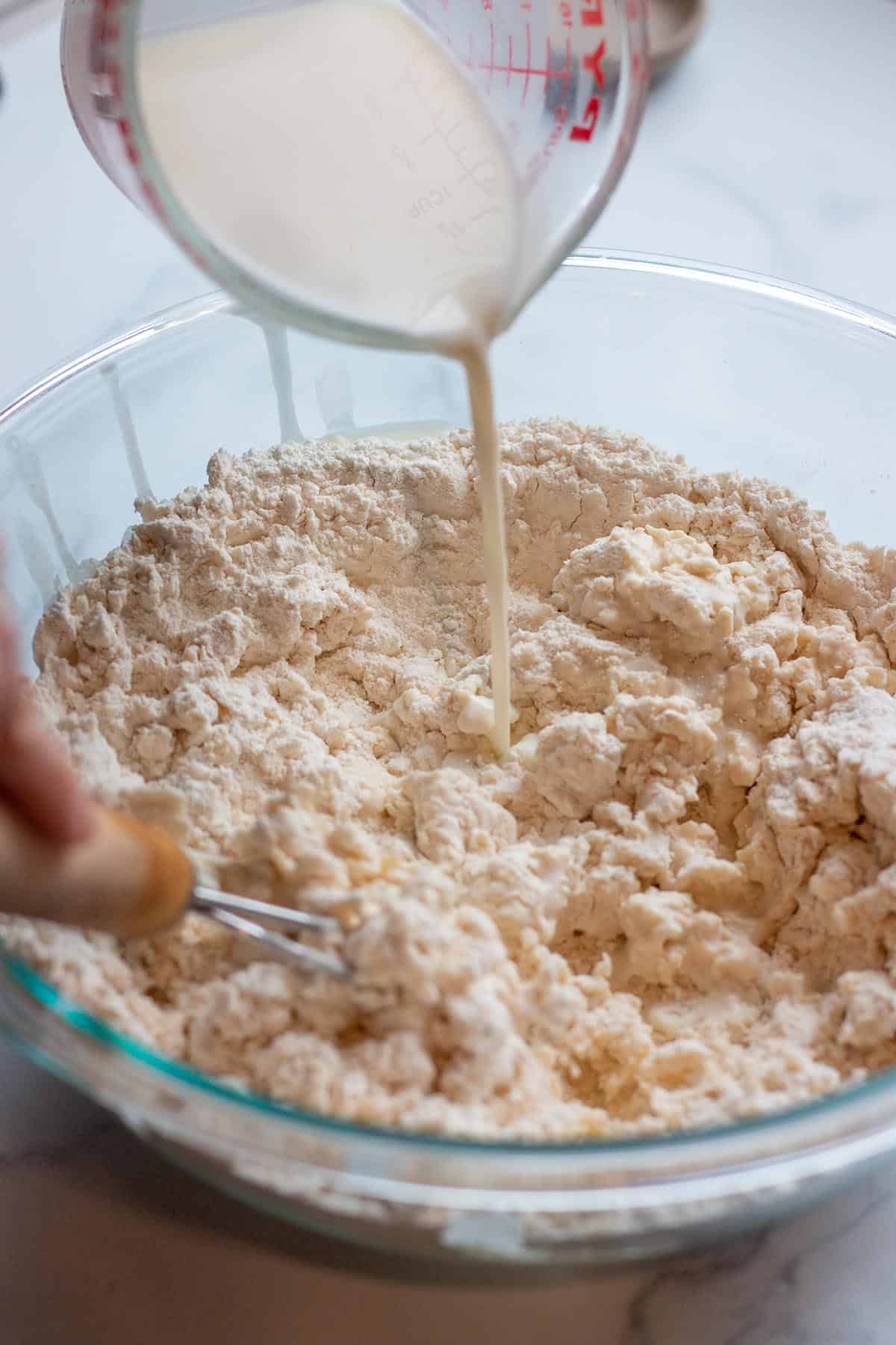 Bowl with flour mix with milk and vinegar being poured in. A danish dough whisk mixing everything together. 