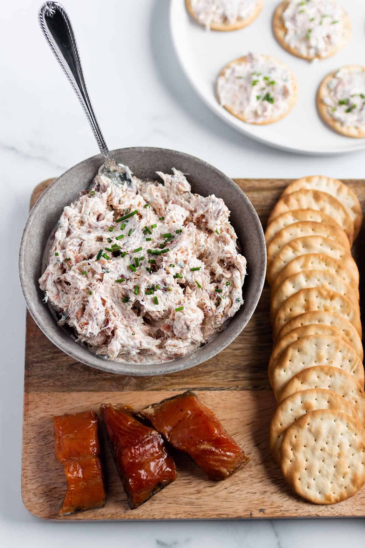 A bowl with smoked salmon dip on a wooden board with crackers and pieces of smoked salmon. 