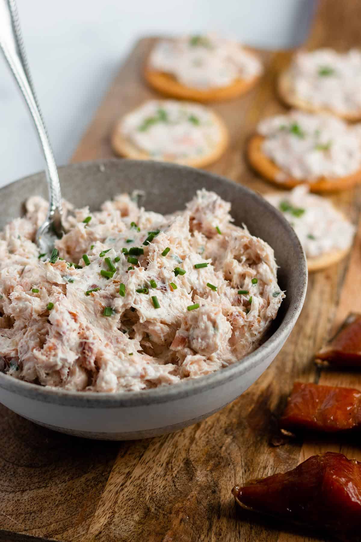 A bowl of smoked salmon dip topped with freshly cut chives with crackers and smoked salmon spread and pieces of smoked salmon. All on a wooden board. 