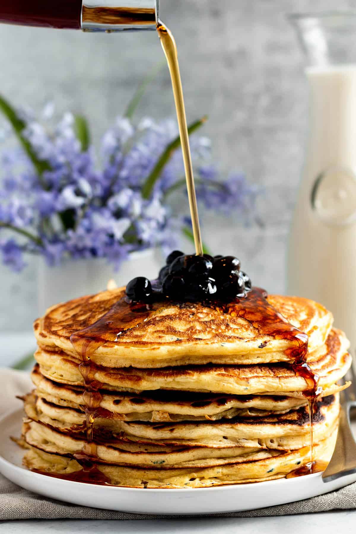 A tall stack of sourdough pancakes with blueberries on top and syrup being drizzled.
