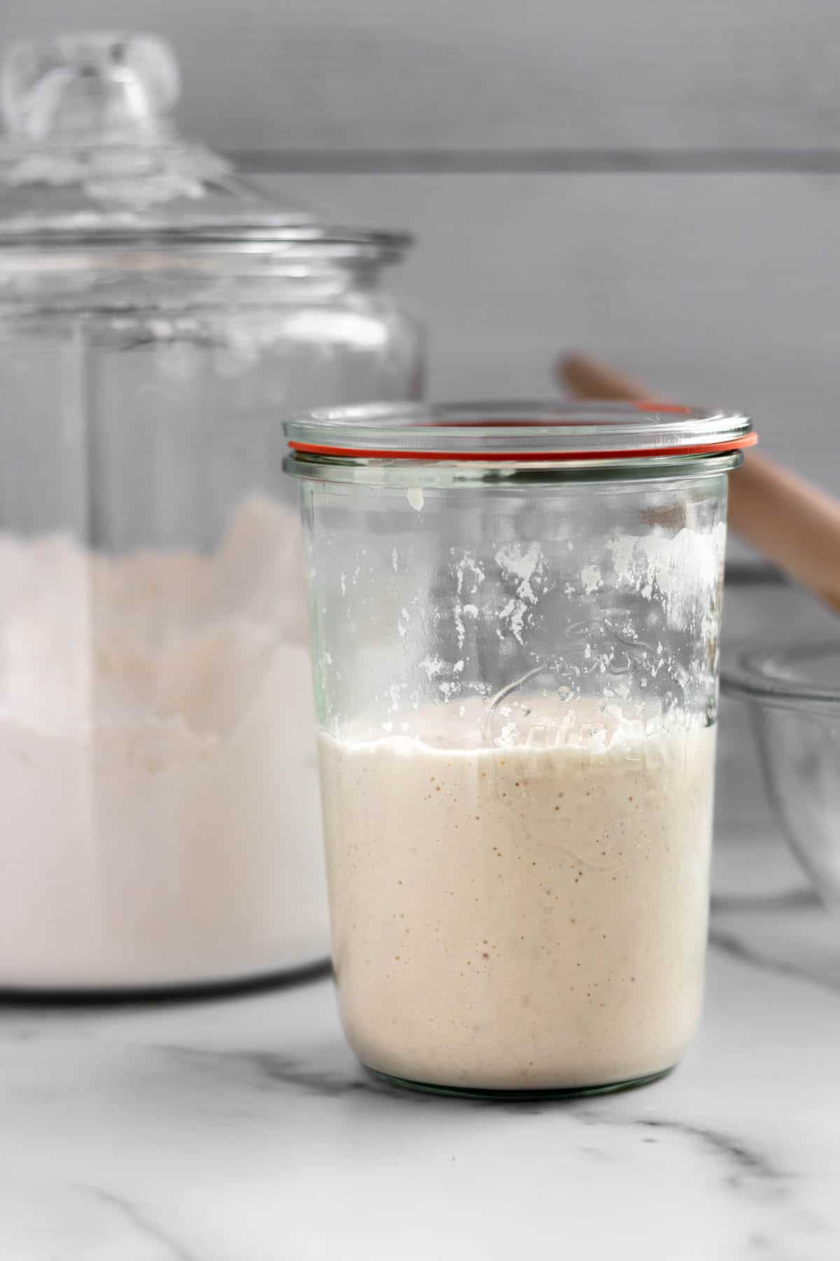 A glass jar with sourdough starter, flour and a dough whisk.