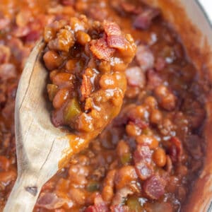 A large skillet with baked beans and a wooden spoon taking a scoop out.