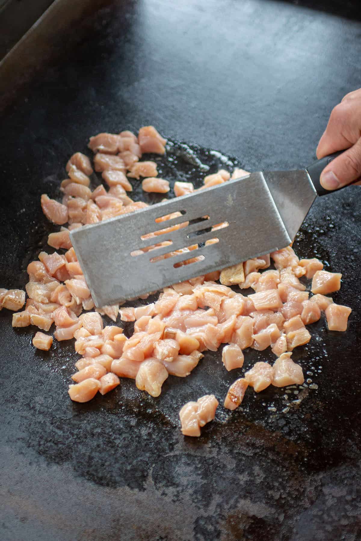 Diced chicken being cooked on a Blackstone with a metal spatula. 