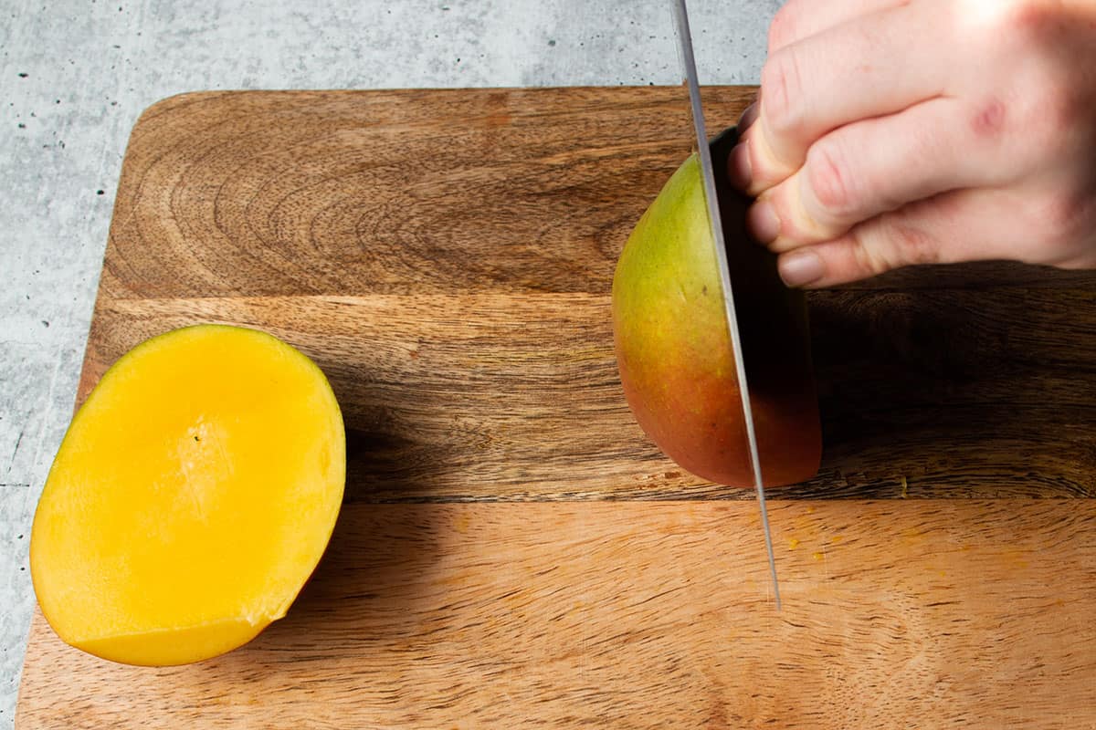 A knife cutting the second side of a mango with the fist side removed.