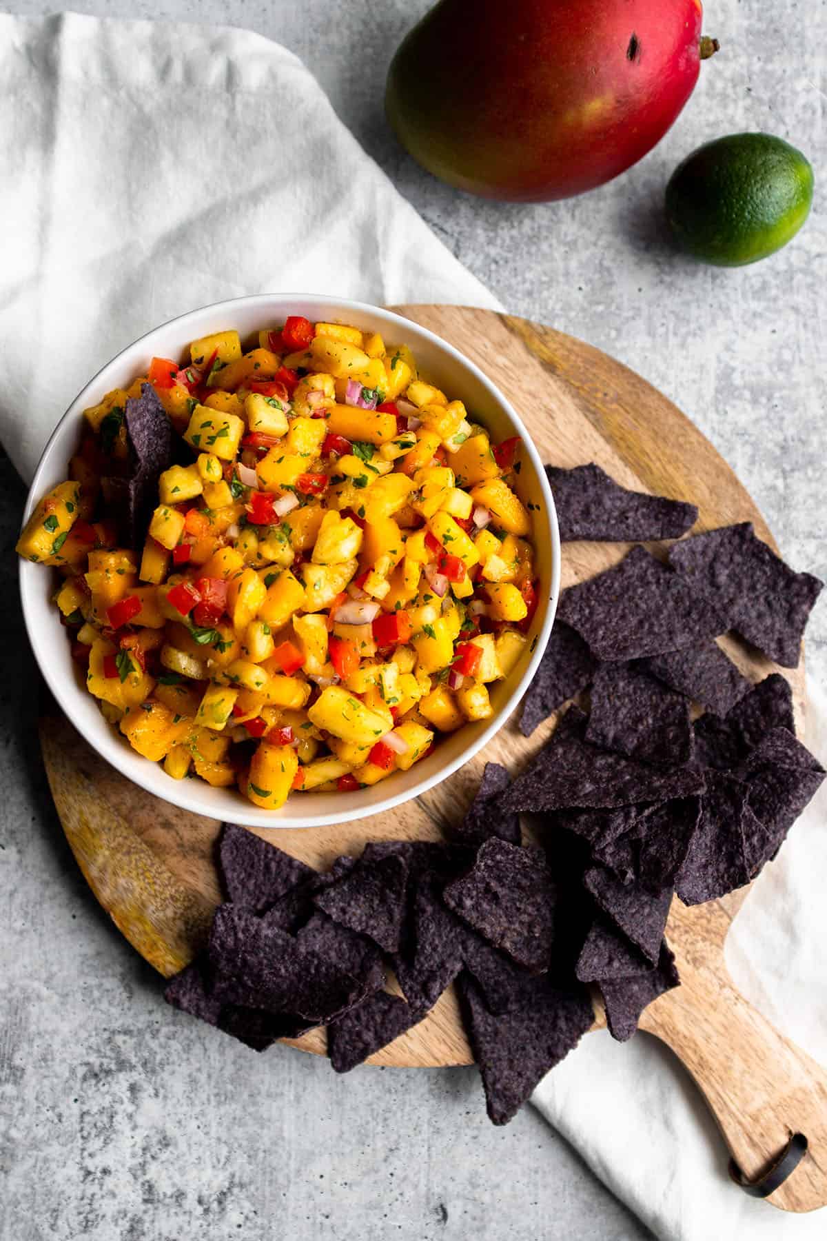 A circle wooden board with a bowl of mango salsa and blue corn tortilla chips with a fresh mango and lime.