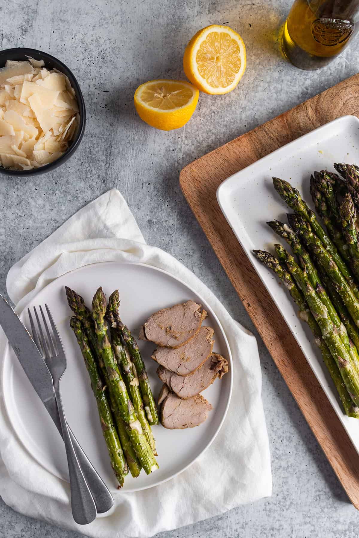 Plate with asparagus and pork with a cutting board with more asparagus, a bowl of parmesan, lemons and olive oil. 