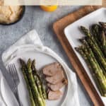 A plate with asparagus and pork with a cutting board with more asparagus.