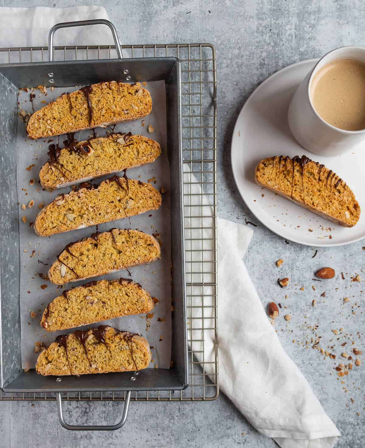 A wire cooling rack with a metal serving tray with almond and chocolate biscotti with a plate and mug of coffee on the side. 