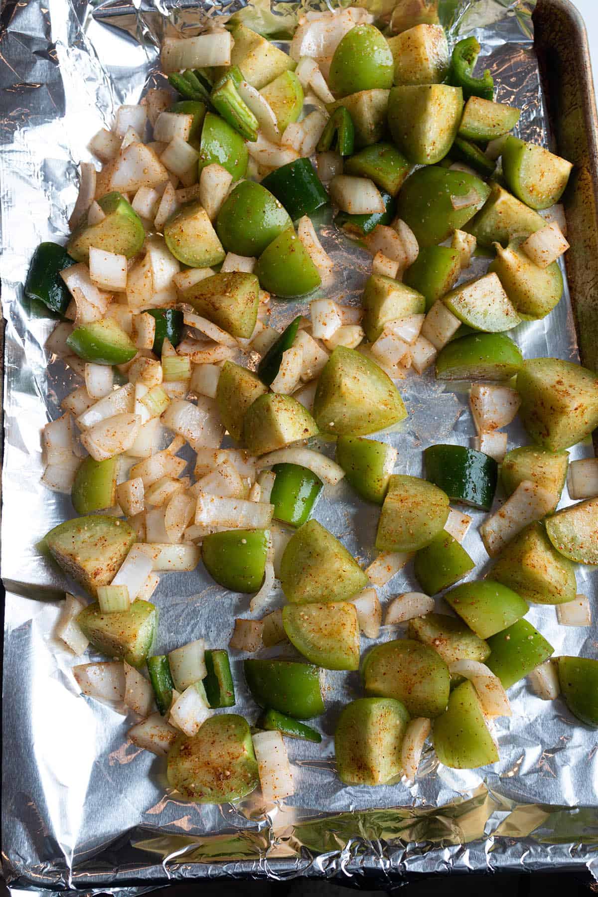 A rimmed baking sheet with aluminum foil and a single layer of tomatillos and onions. 